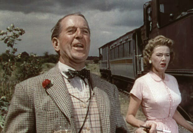 An older man well dressed bowie with a younger woman stands next to a train looks happy