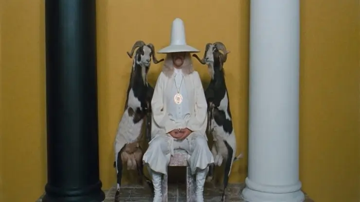 A man in a white hat and clothes sits down between 2 goats