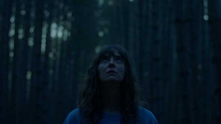 a dark haired woman stands looking up in a forest
