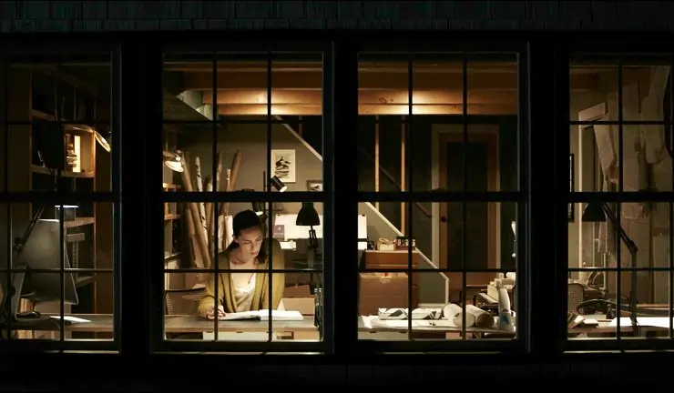 A woman studies at night in a house with large windows in The Night House.