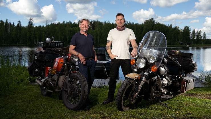 two men stand next to their bikes at a lake