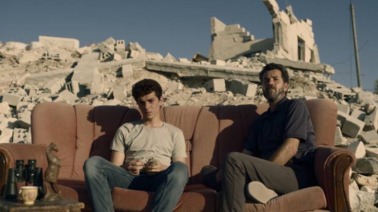 A man and young man sit on a sofa in war torn land