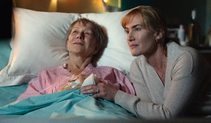 An older woman lies on a bed with a younger woman holding her hand