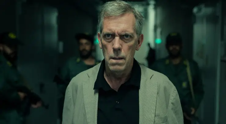 An older man looks concerned with armed soldiers behind him (Hugh Laurie in Tehran Season 3).