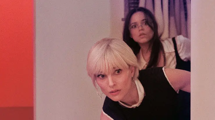 Blonde haired woman sneaks to look around as a dark haired woman behind her in The Gallerist.