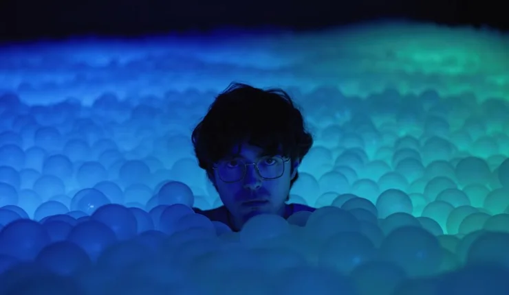 A dark haired young man (James Sweeney) in glasses sits in the middle of glowing balls in Twinless.