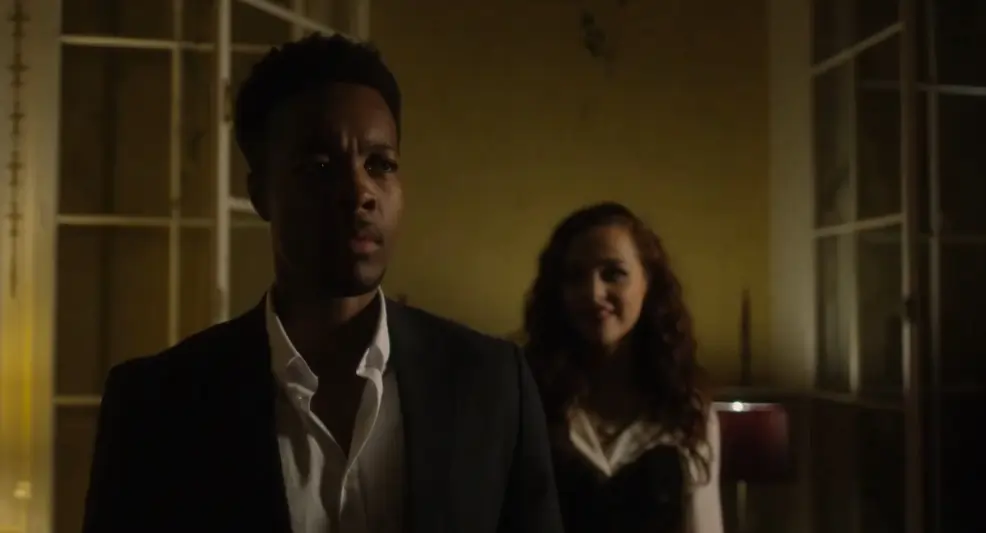 A man stands in dimly lit room with a woman behind him in Lure