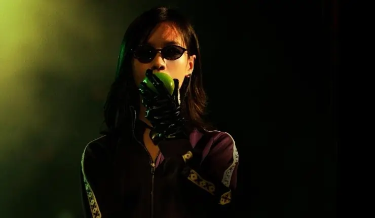 A Asian stands in the dark with sunglasses eating an apple.