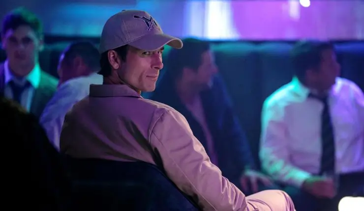 A man in a cap and matching jacket sit in a night club watching someone