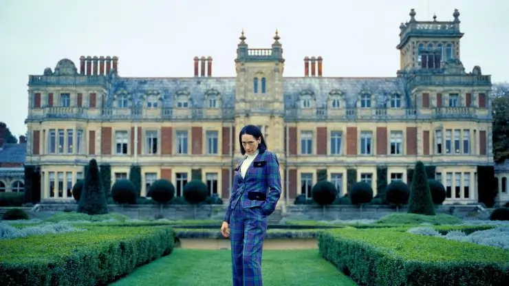 An middle aged woman stands in the gardens of a manor. in Lady