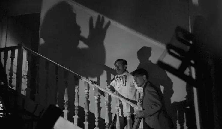 2 men stand on staircase in fear as a dark shadow lurks over them in The Devil's Hand
