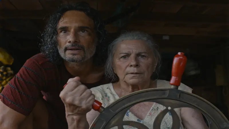 An older woman stirs a wheel on ship with a younger dark haired man behind her in Blue Trail