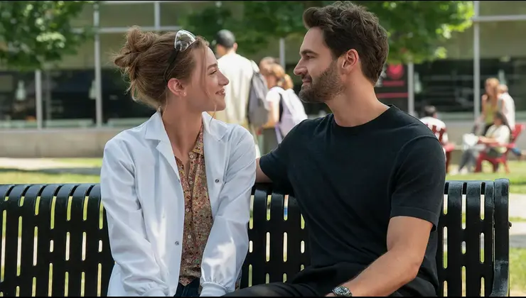 Lili Reinhart and Tom Bateman sit on a bench outdoors, sharing a warm, close moment as they look at each other in a scene from The Love Hypothesis.