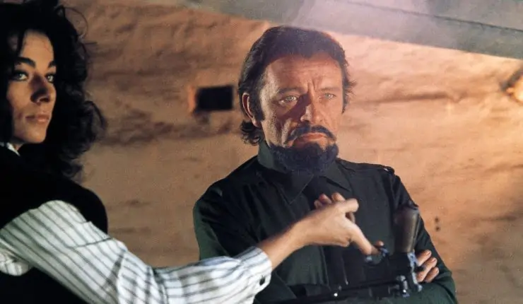 A dark haired and beard man is hand a gun by a woman in Bluebeard (1972)