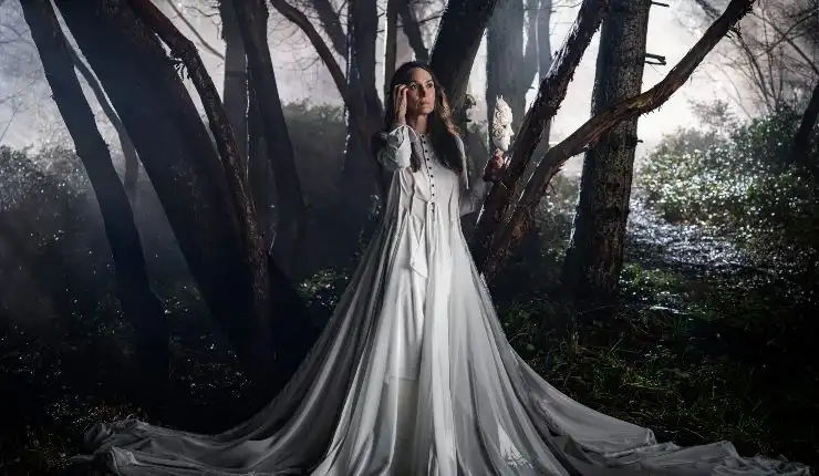 An older woman wears a flowing silver dress in a dark forest.