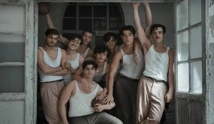 A group of seven young men in white vests and trousers pose together in the doorway of a weathered building, with one holding a basketball.