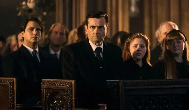 A man suited in black sits with others at a funeral in Rivals Season 2