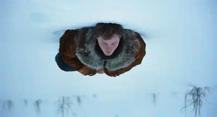 A younger man in a fur coat lies in the snow in A Long Winter