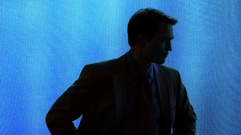 A man in a suit stands in the shadows in front of a blue background.