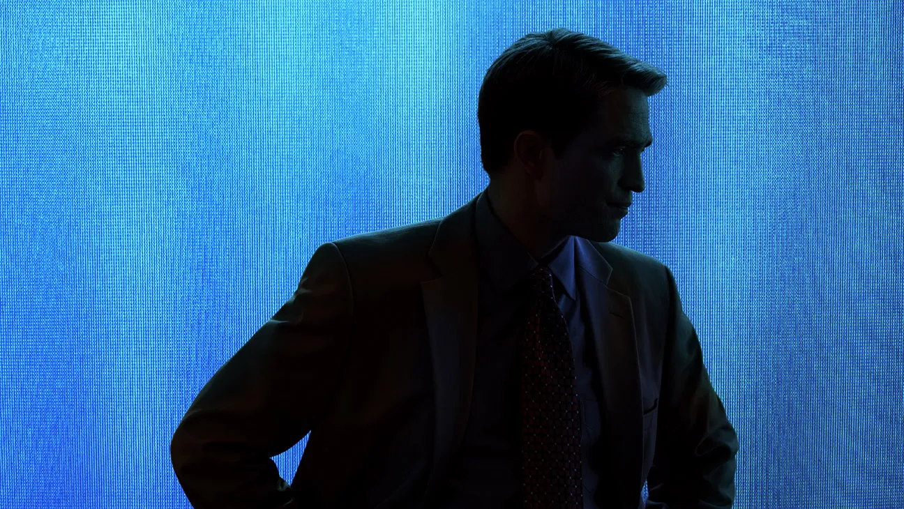 A man in a suit stands in the shadows in front of a blue background.